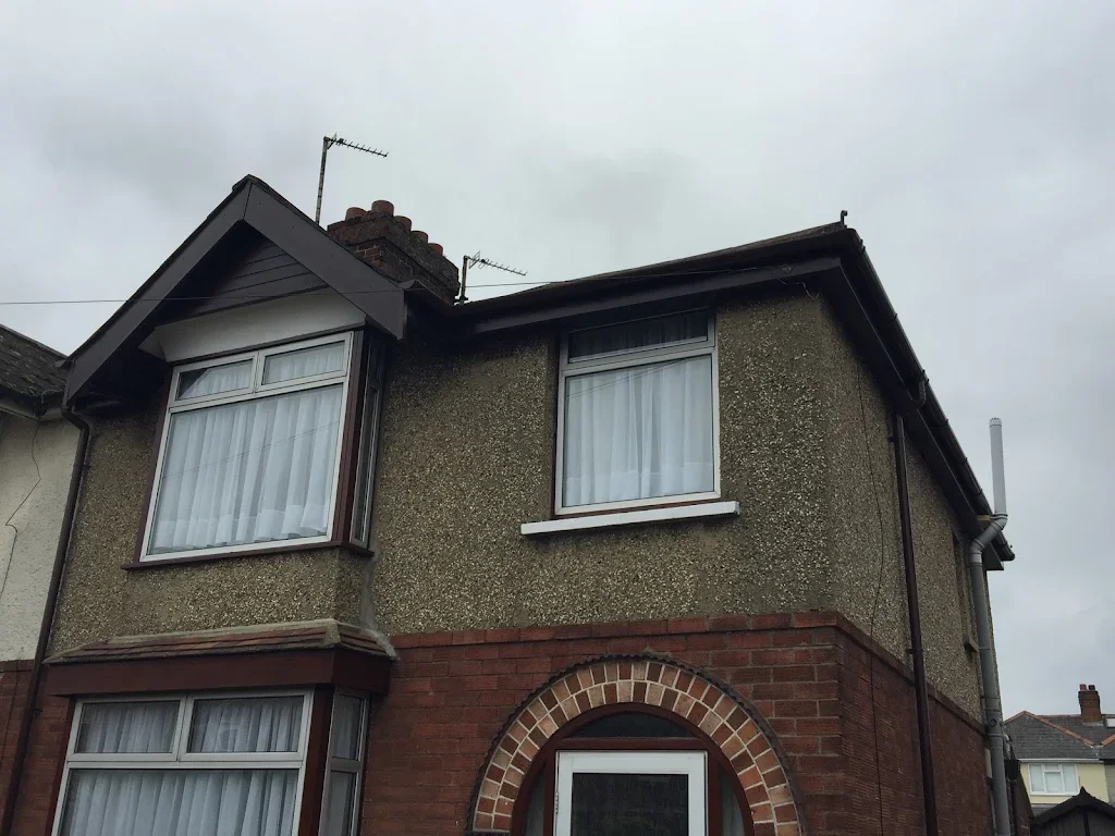 Front of a two-story house with brick arch around the door and two large windows on the upper floor, with curtains inside. The house has a textured exterior with a section of brickwork at the bottom and is under an overcast sky.