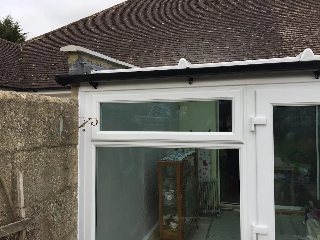 Close-up of a modern white patio door or window with a black gutter, set into a house with a tiled roof, next to a concrete wall with a rusty metal hook.