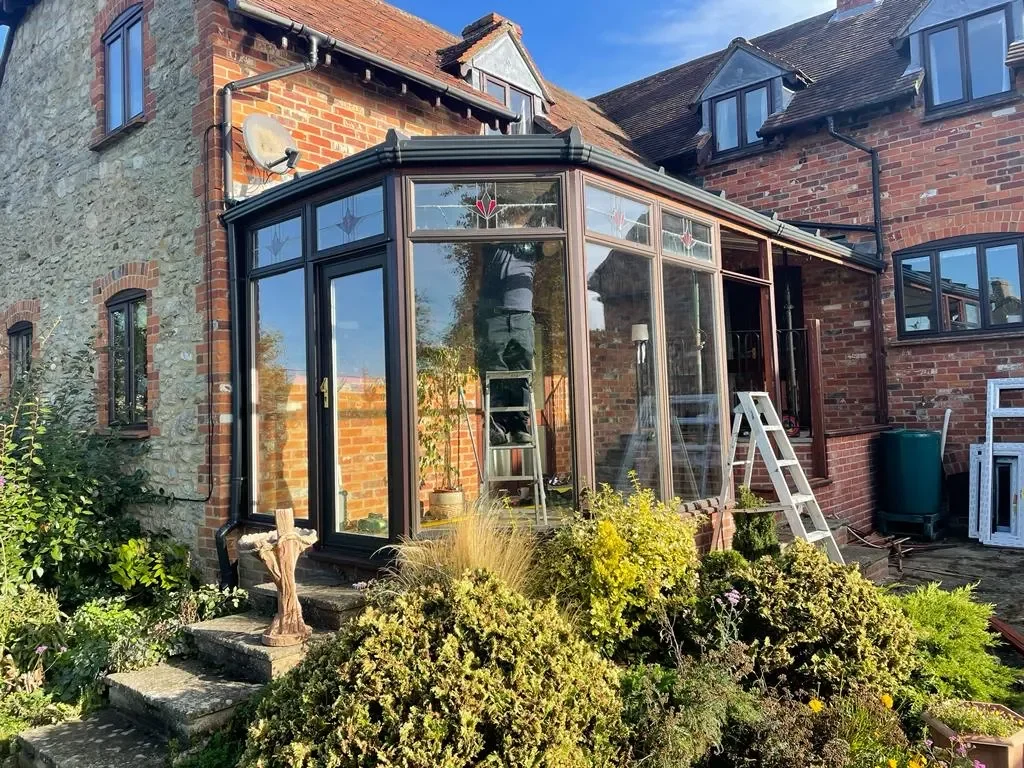 A brick house with a glass enclosure extension, with ladders inside and outside, and garden plants and steps in the front.