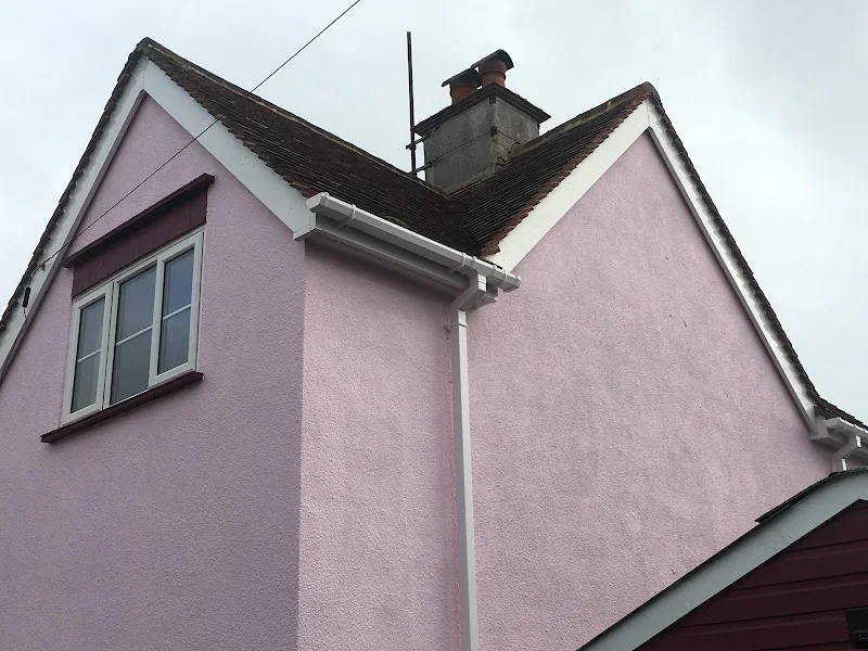 Close-up of a pink house with white trim, showing a window and roof gables, with a chimney and overcast sky in the background.