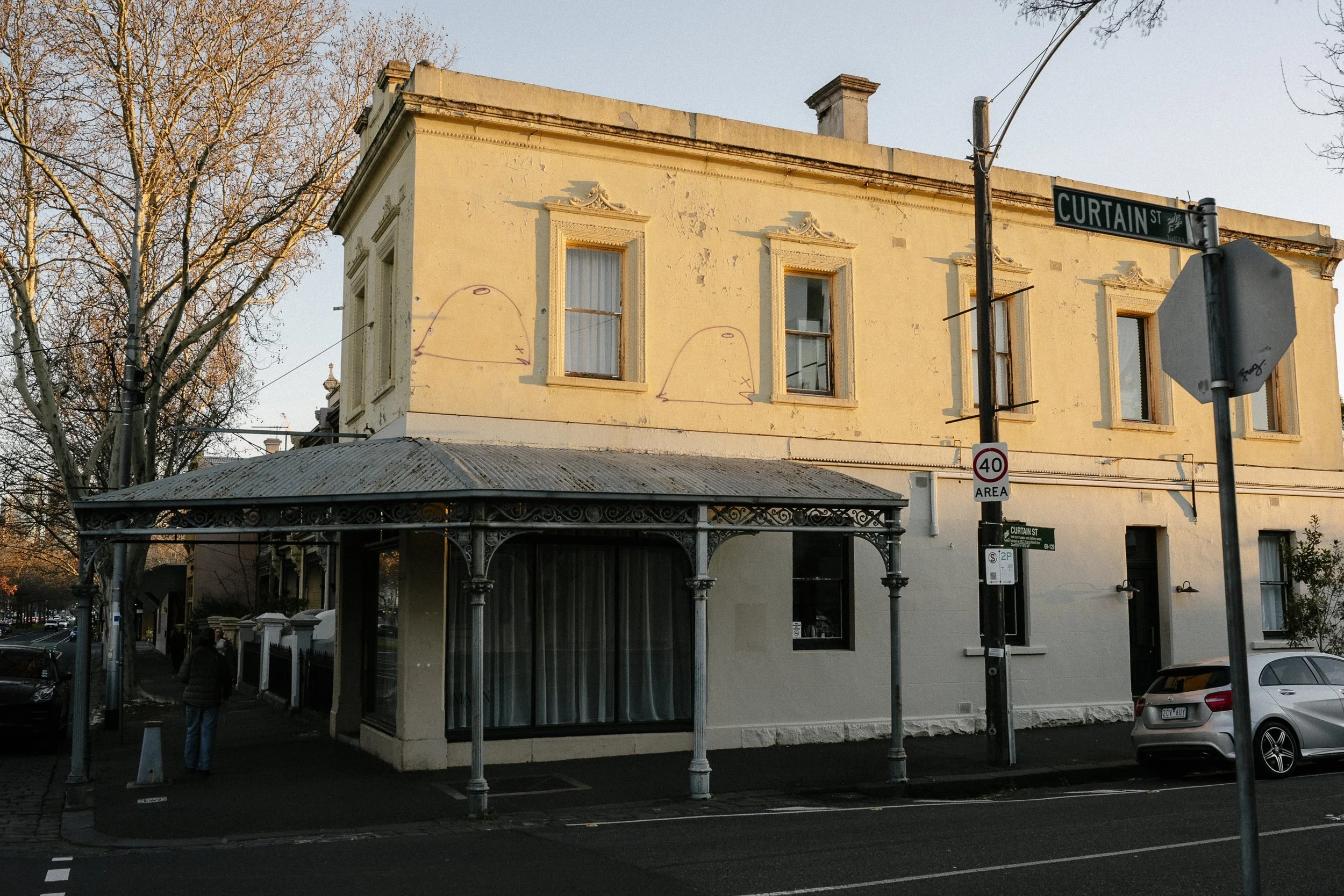 A corner building with a beige exterior, three windows, and an ornate iron awning covering the sidewalk at the corner of Curtain Street and Rathdowne St, Carlton North.
