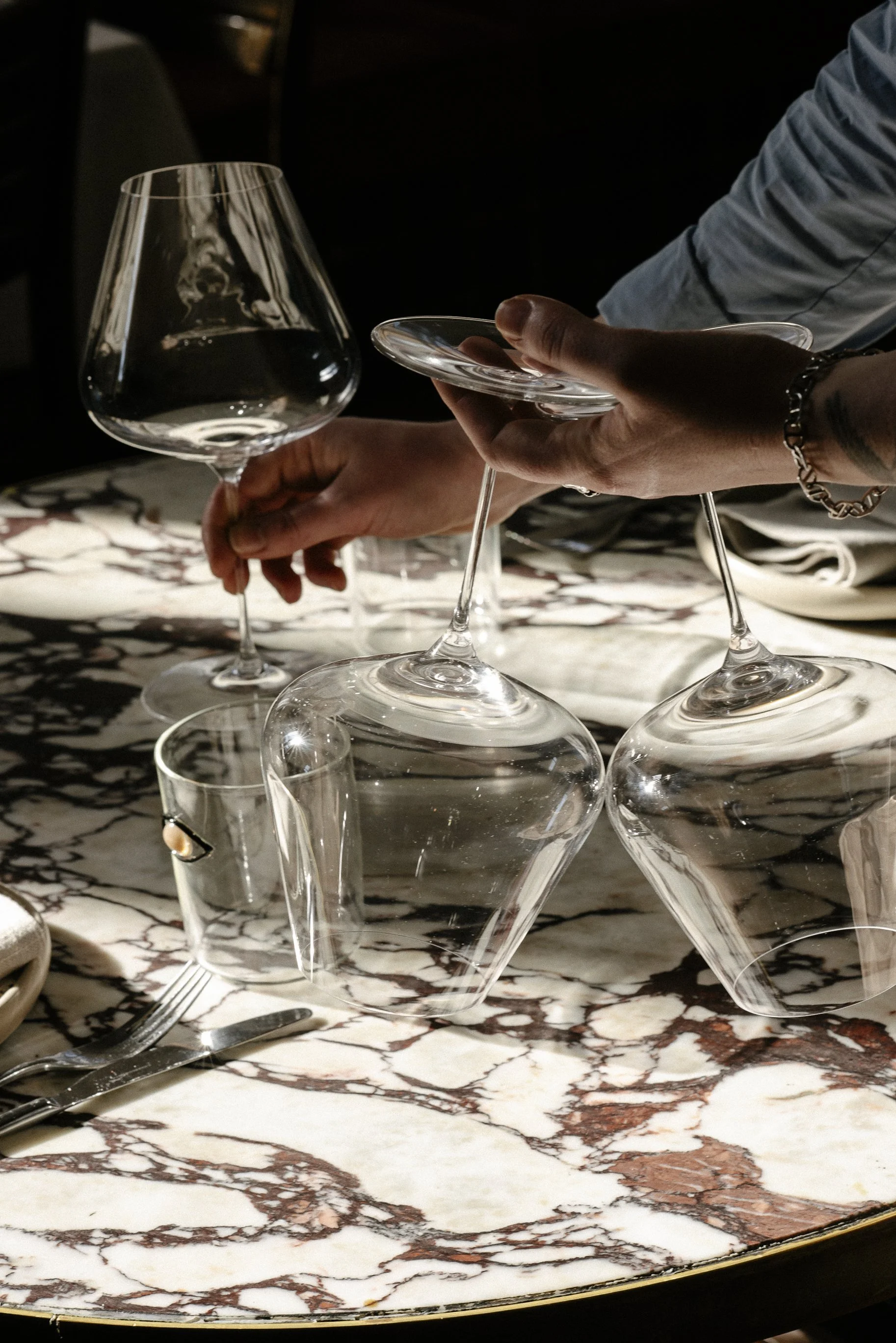 A person arranging empty wine glasses on a marble table, with silverware nearby.