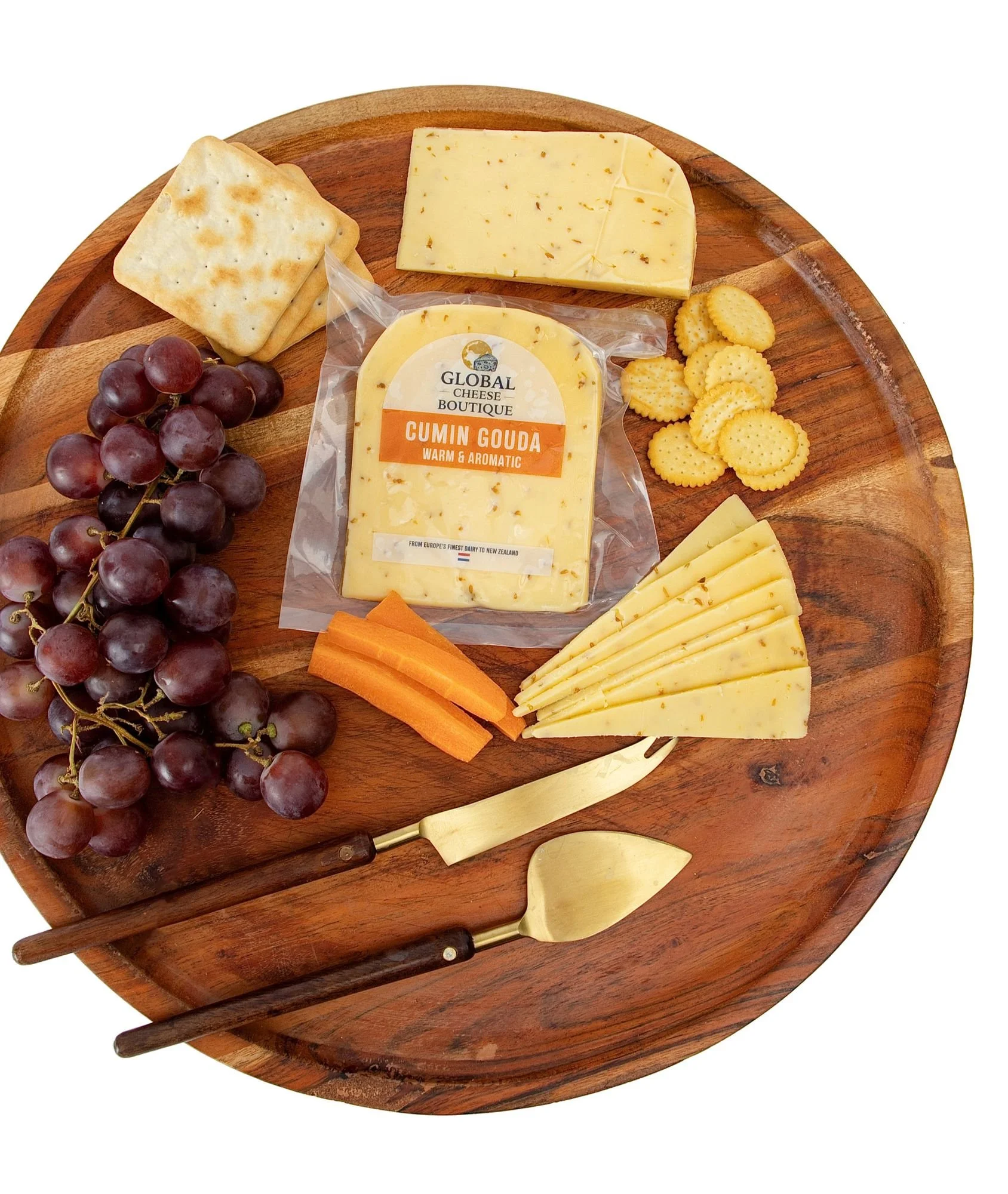 Cumin Gouda sealed pack with a wedge, slices, grapes, crackers and carrot sticks on a wooden board
