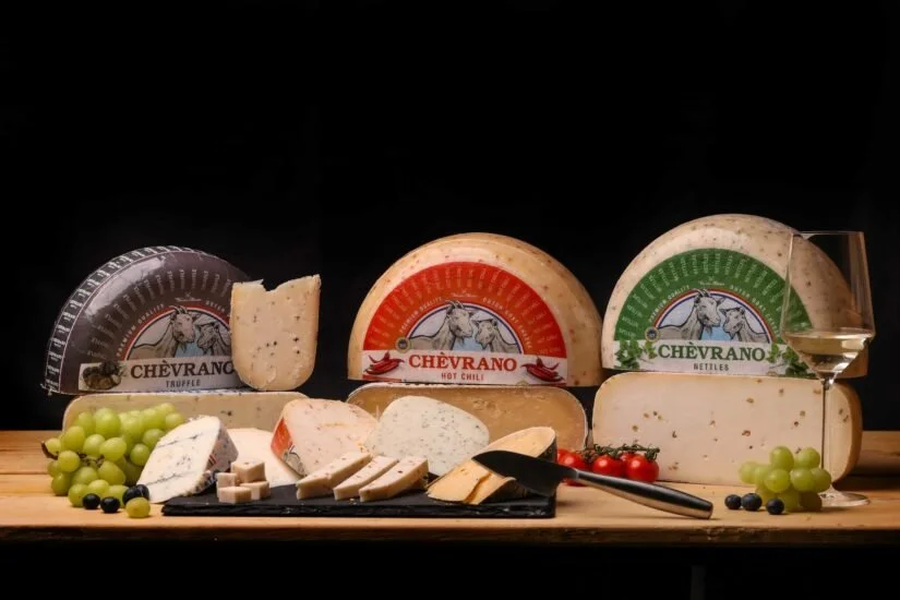 Display of various cheeses, grapes, cherry tomatoes, and a glass of white wine on a wooden board against a black background.