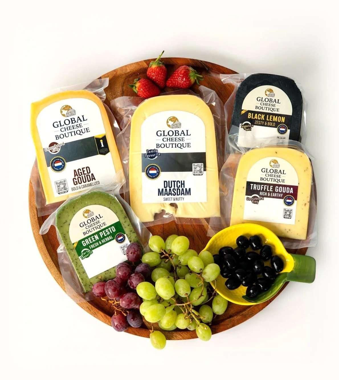 Build Your Own 5 example: Aged Gouda, Dutch Maasdam, Black Lemon, Truffle Gouda and Green Pesto packs on a wooden board with grapes, strawberries and olives
