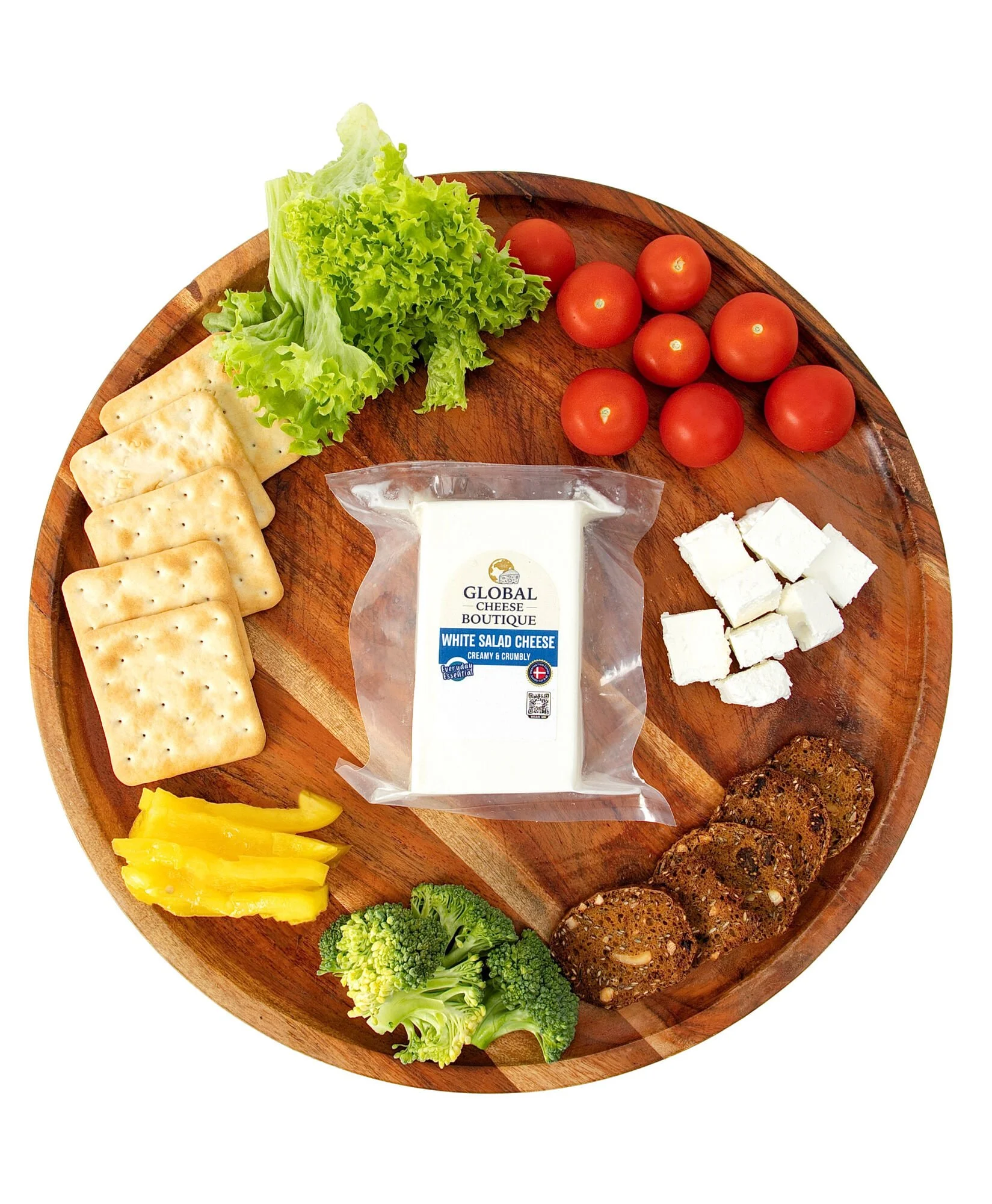 Overhead view of White Salad Cheese 230 g on a round board with cubes, lettuce, cherry tomatoes, crackers, capsicum and broccoli
