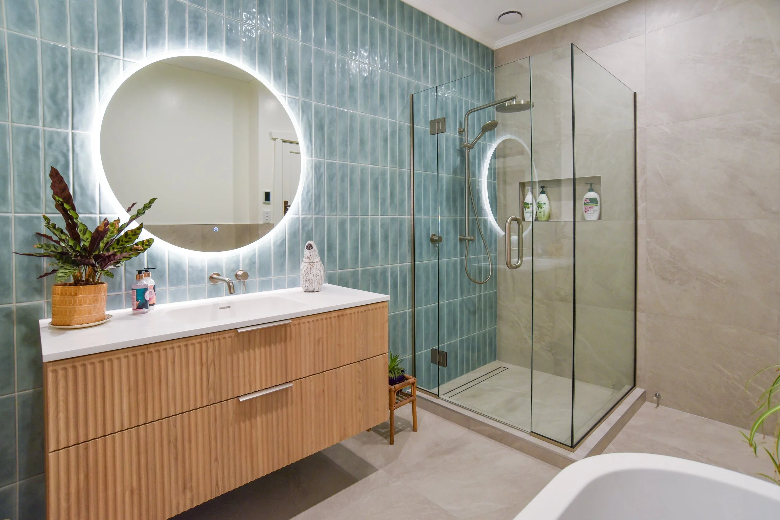 Modern bathroom with a backlit round mirror, a white sink with a wooden vanity, a potted plant, and a glass-enclosed shower with blue tiles and built-in shelves.