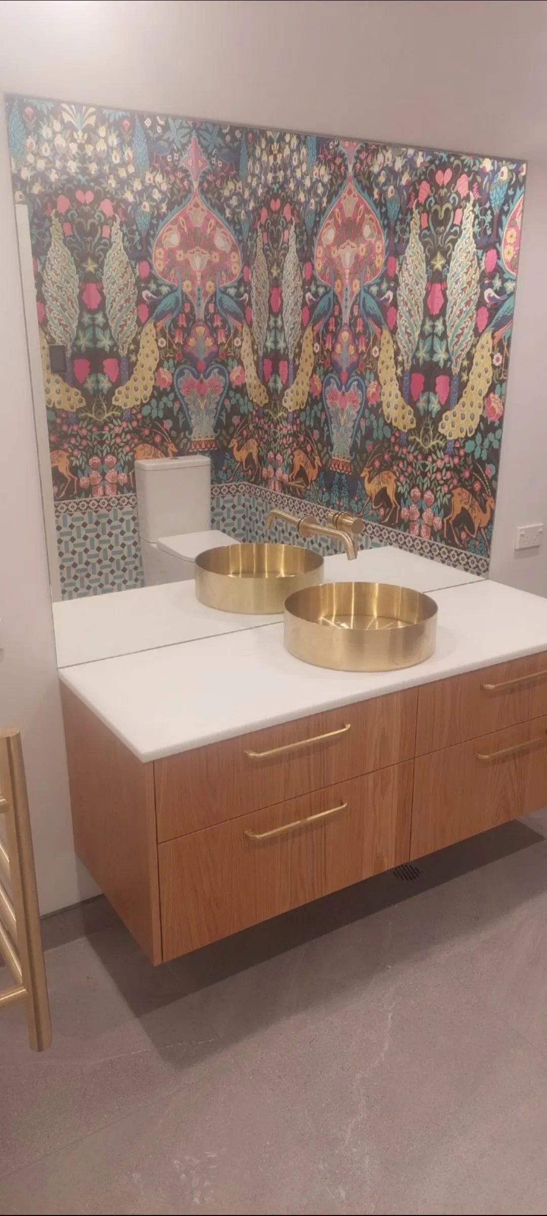 Bathroom vanity with a large mirror, two round gold sinks, and gold faucet, with colorful, patterned tiles on the wall and a wooden cabinet below.