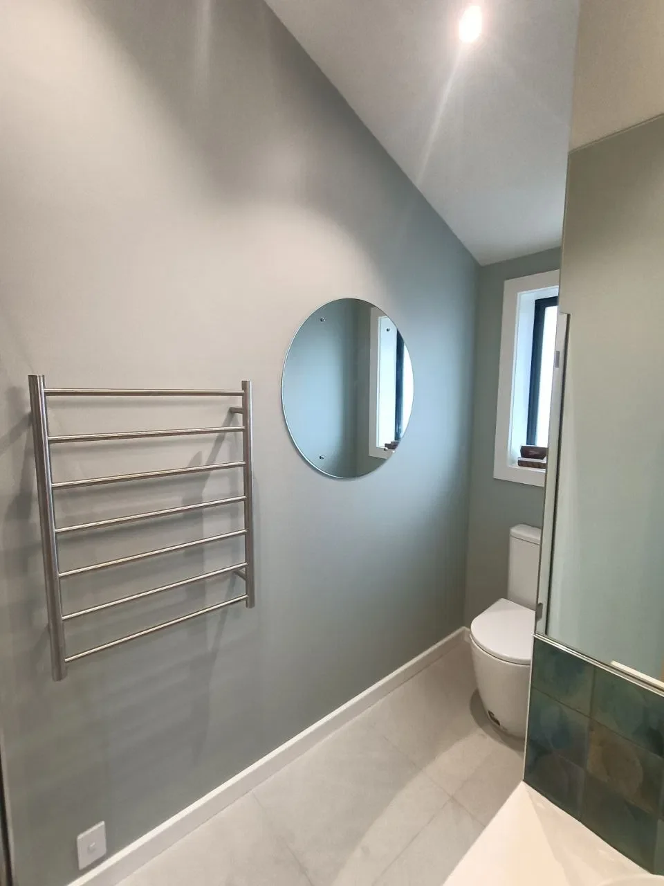 Modern bathroom with a gray wall, a round mirror, a small window, a white toilet, and a metal towel radiator.