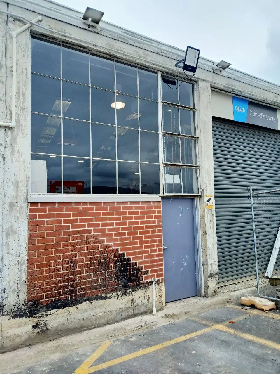 Exterior of an old industrial building with large window panes, a brick wall section, a gray metal door, and a rolled shutter door, with a sign reading "Dunedin Store" and a blue "Delta" sign.