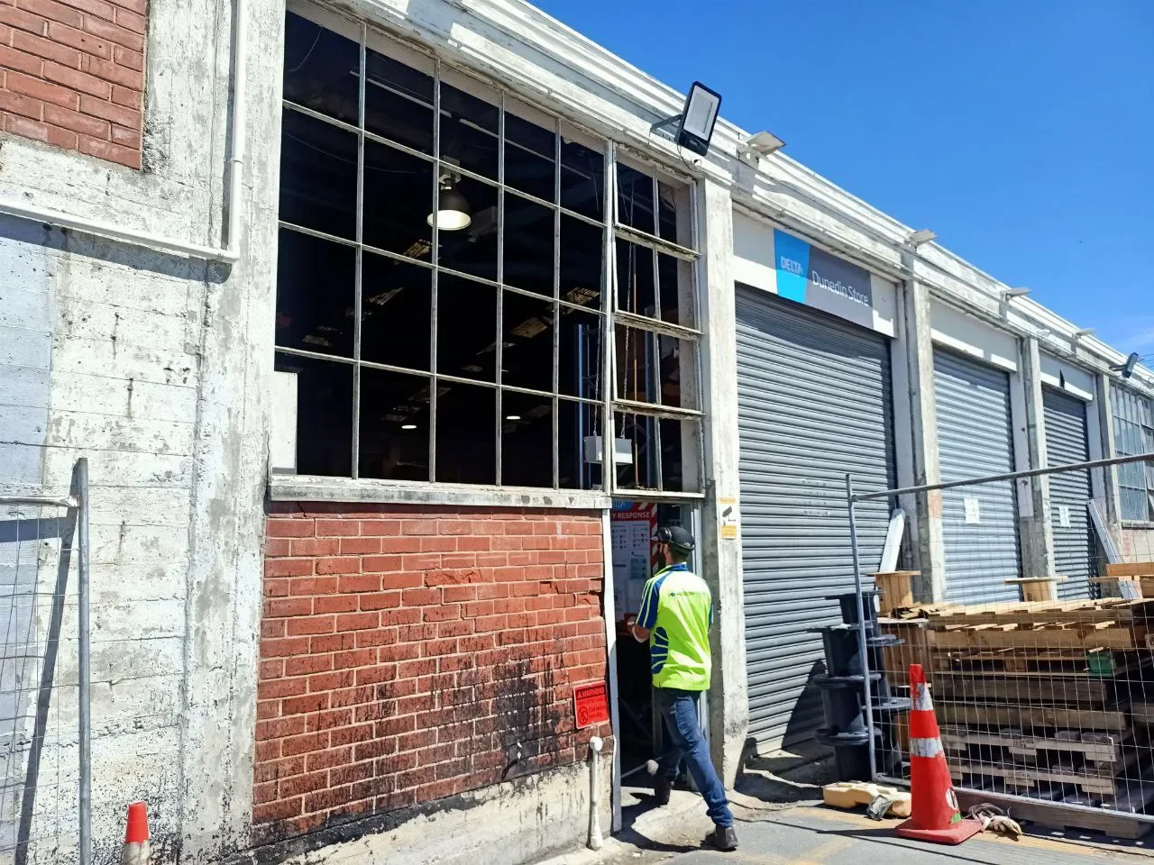 A construction site with a person in a yellow safety vest and hard hat standing near a partially opened shutter door. The building has large windows, some of which are missing glass panes, revealing the interior. There are orange traffic cones and co