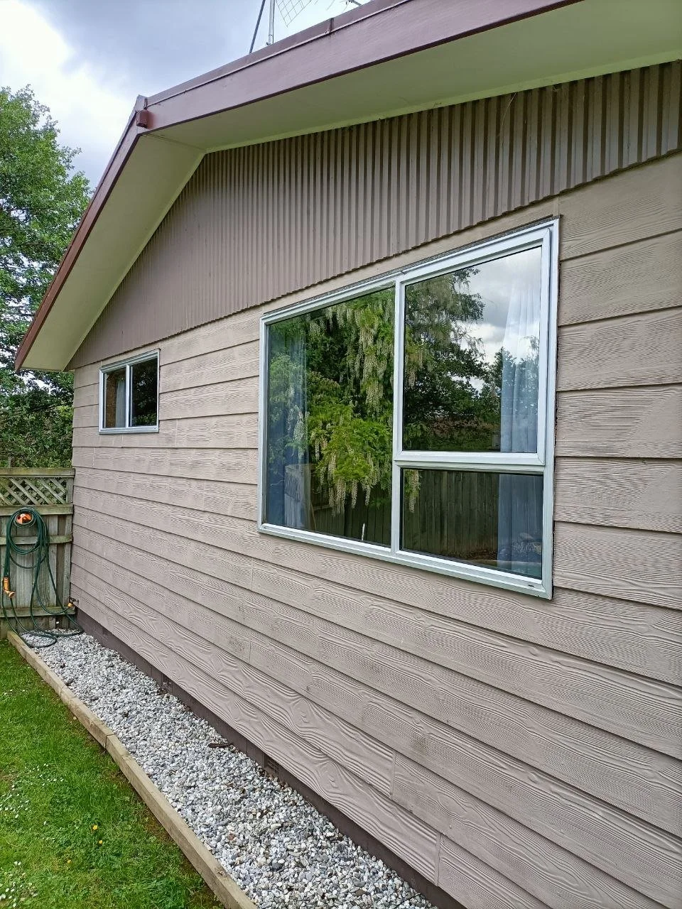 Exterior of a house with horizontal beige siding, a large window with white framing, and a smaller window, with a gravel bed along the foundation and a garden hose attached to a wall-mounted reel.