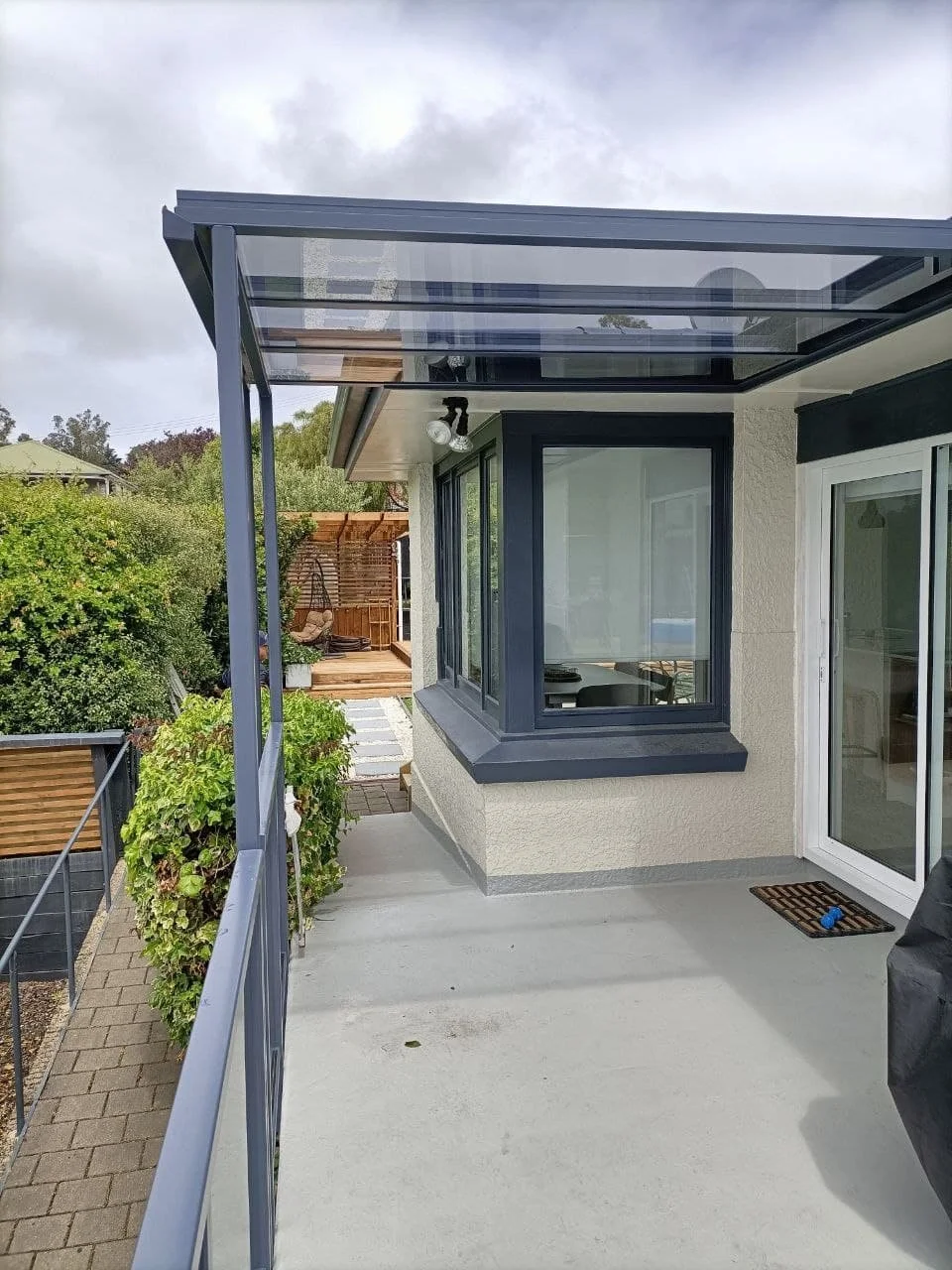 A small outdoor patio area attached to a house with a glass roof, black window frames, a beige exterior wall, and a white sliding door. There's a metal railing along the edge of the patio and greenery in the background.