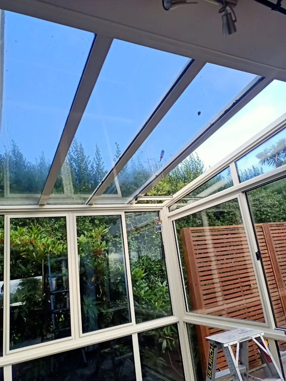 A glass conservatory with a clear glass ceiling and walls, surrounded by lush green plants and trees outside, with a wooden privacy screen on one side and a stepladder inside.