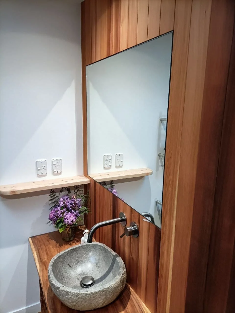 Bathroom with a stone sink, wooden countertop, large mirror, purple flowers in a vase, and wood-paneled wall.