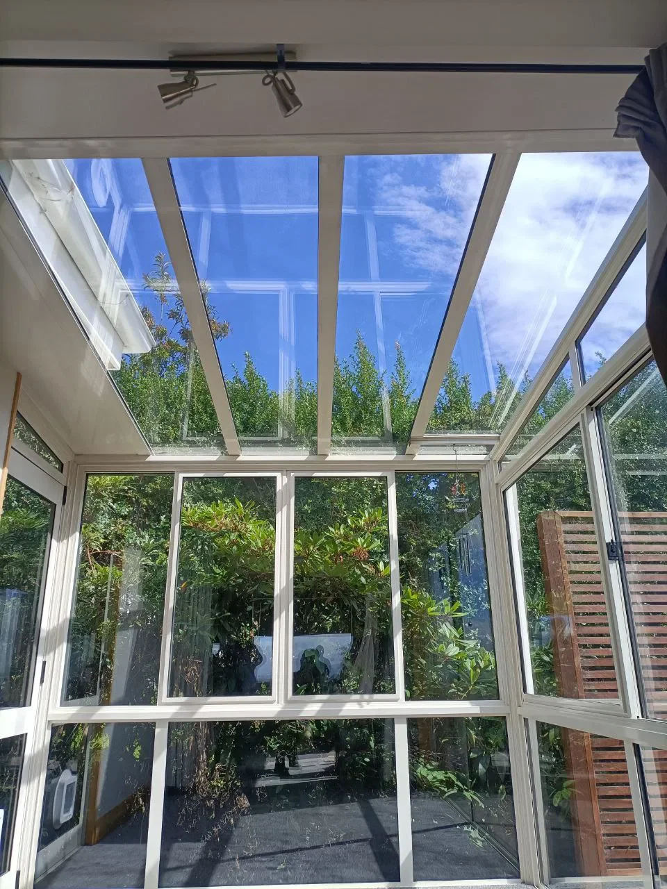 A glass conservatory with white frames, showing a view of green foliage outside and a cloudy blue sky above.