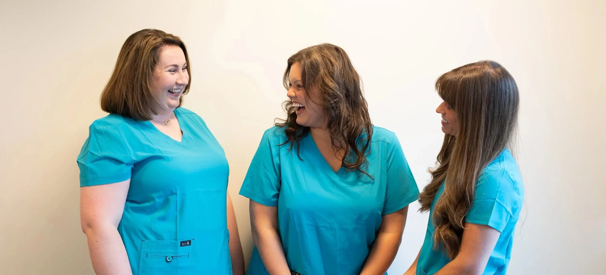 Three women wearing blue medical scrubs smiling and laughing together in front of a plain beige wall.