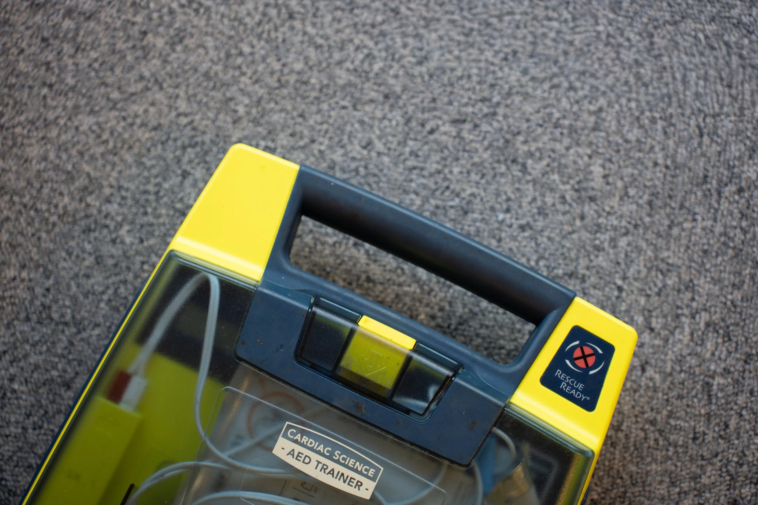 Top view of a yellow and black AED trainer with a handle, placed on a gray carpeted surface, labeled 'Cardiac Science AED Trainer'.