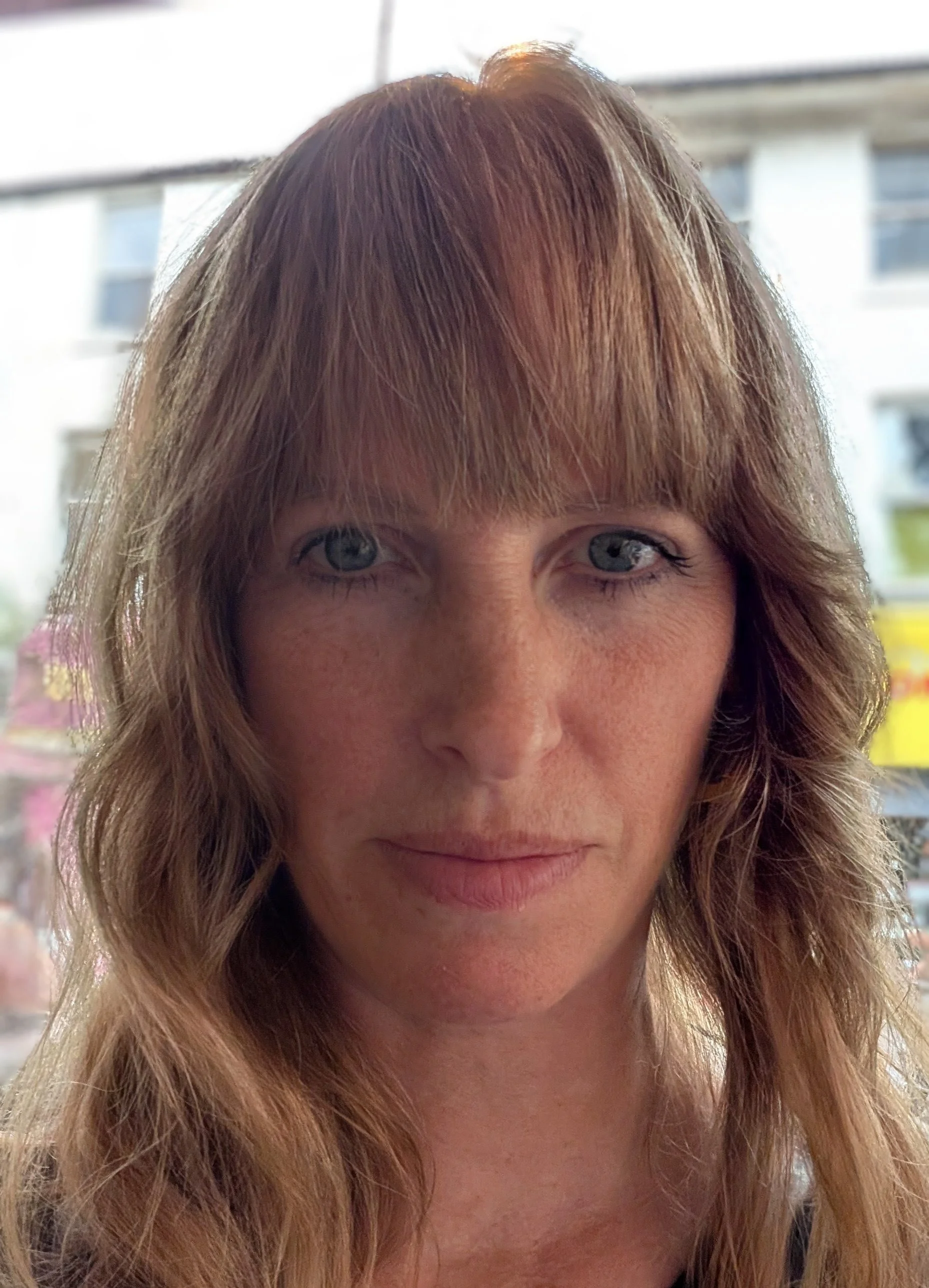 Close-up of a woman with light brown hair and blue eyes indoors, with a background of window and building outside.