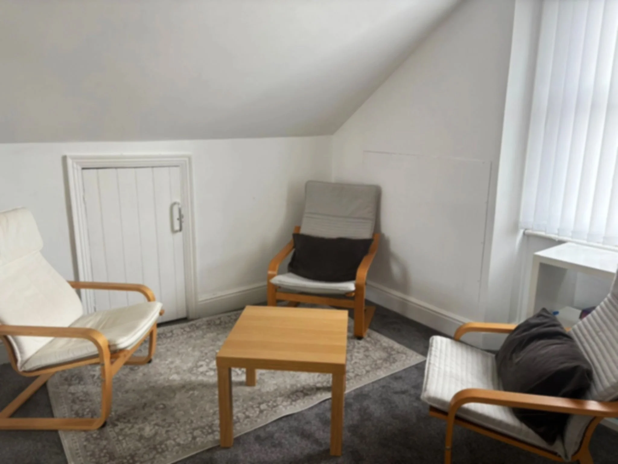 A cozy therapy room with three wooden armchairs, a small wooden table, a patterned rug, and a small window with vertical blinds.