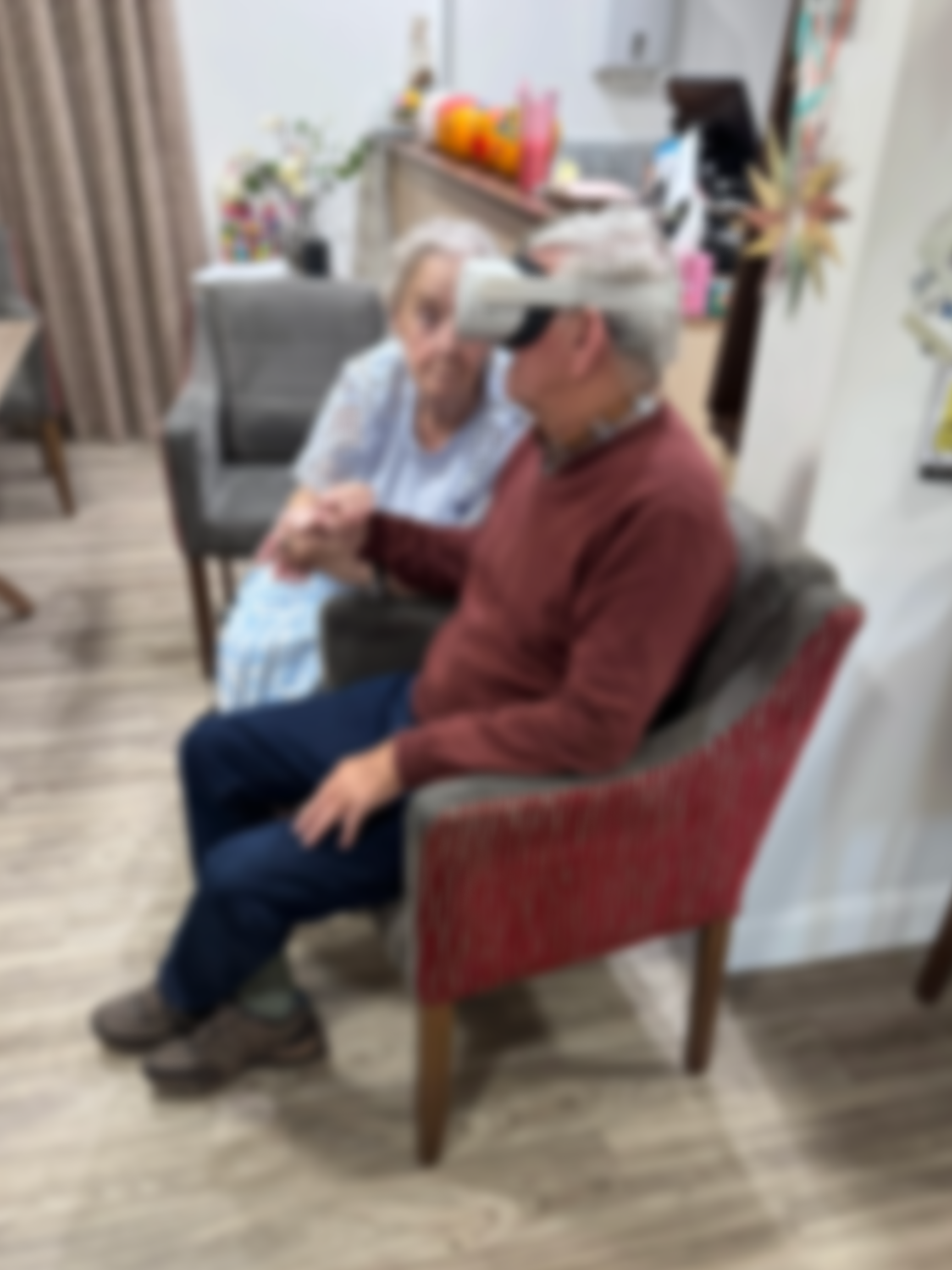 Two elderly people sitting close together in a living room, one with a prosthetic eye, holding hands, with colorful decorations in the background.