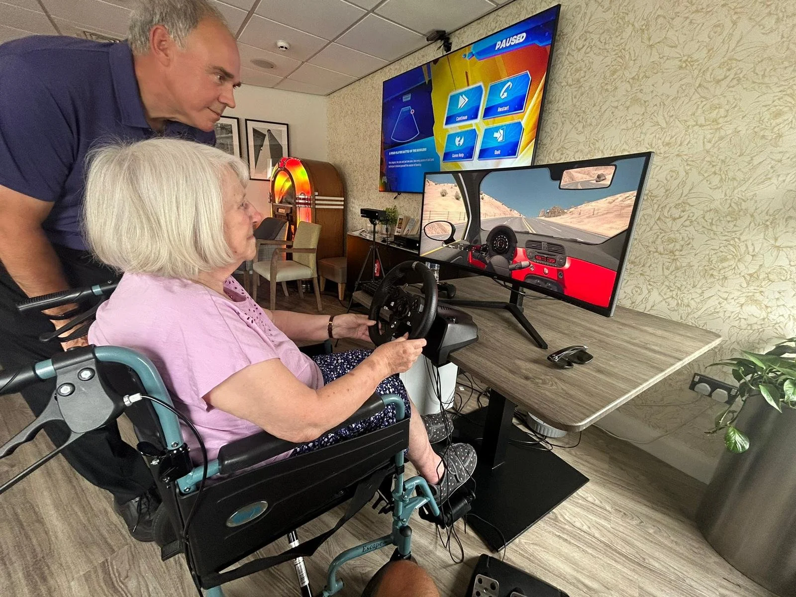An elderly woman in a wheelchair engaging in a driving simulation game on a large monitor, with a man standing beside her observing in a room with arcade machines and framed artwork.