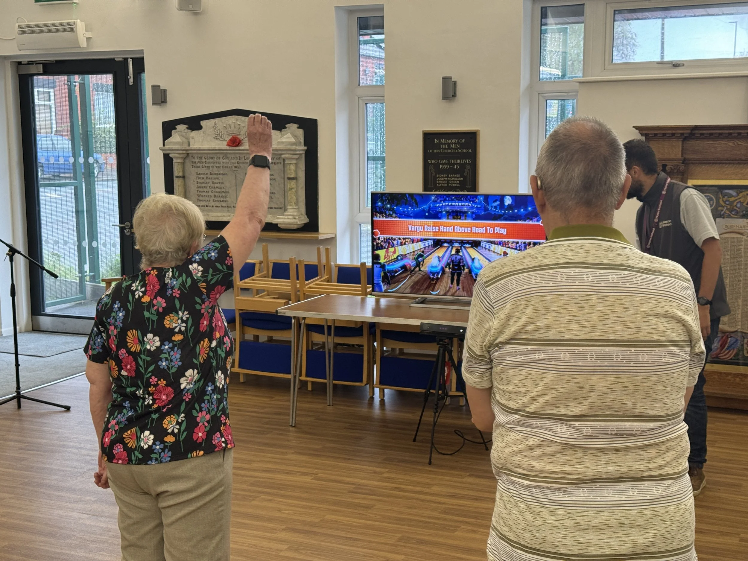 An elderly woman raising her hand in a room watching a demonstration about bowling on a television screen. Two men are also observing, one with his back to the camera and another on the right side. The room has a wooden floor, chairs stacked against the wall, and various plaques and decor.
