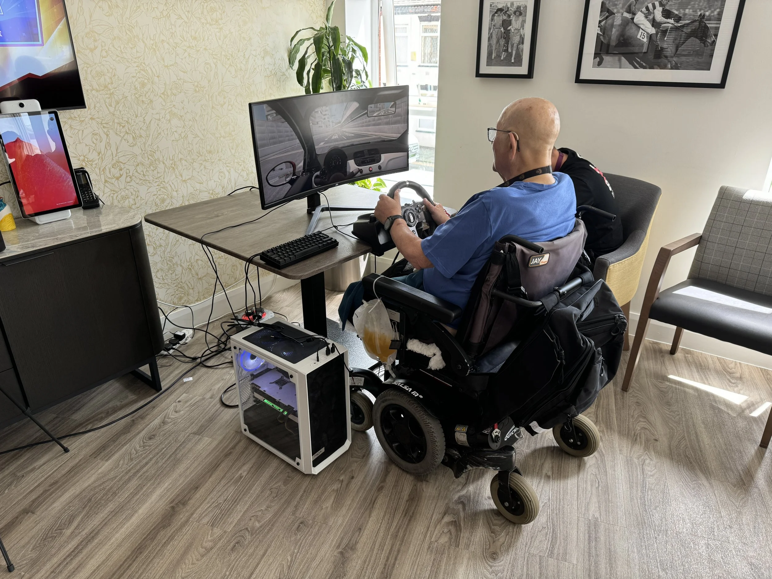 A man in a motorized wheelchair participating in a racing simulation using a large monitor, steering wheel, and gaming setup in a room with wooden flooring, chairs, and framed pictures on the wall.