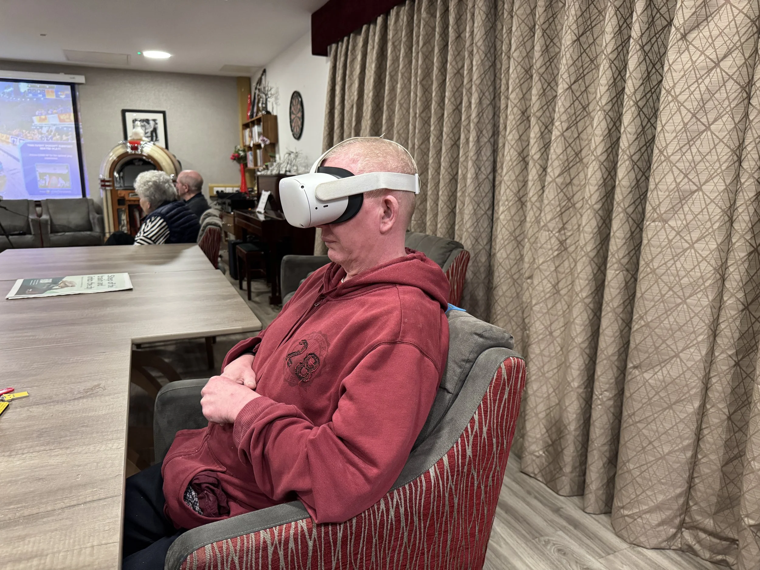 A man wearing a red hoodie and sitting in a red armchair while using a virtual reality headset at a social gathering or party.