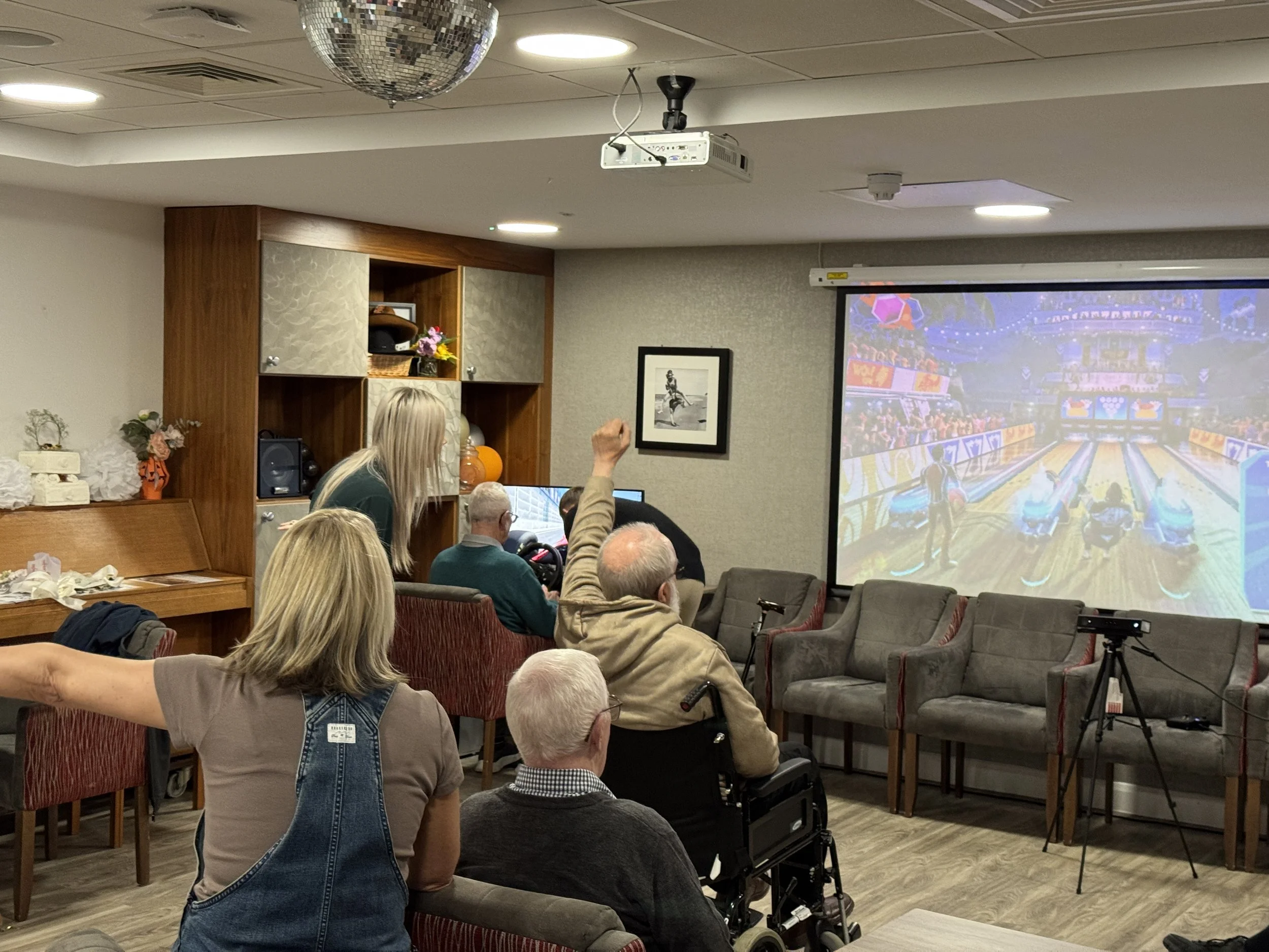 A group of elderly people watching a game of bowling on a large screen in a cozy room, with some seated on chairs and one person in a wheelchair. A woman with blonde hair stands near the screen, pointing at it.