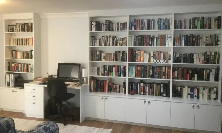A home library with white bookshelves filled with books, a desk with computer and chair, and a small white cabinet on the left.