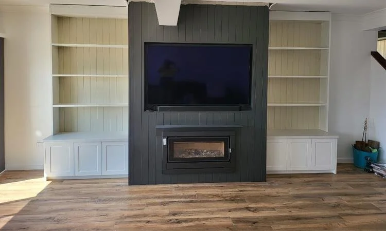 Living room with a flat-screen TV mounted above a fireplace, surrounded by built-in white shelves on either side, hardwood flooring, and a dark accent wall behind the TV.