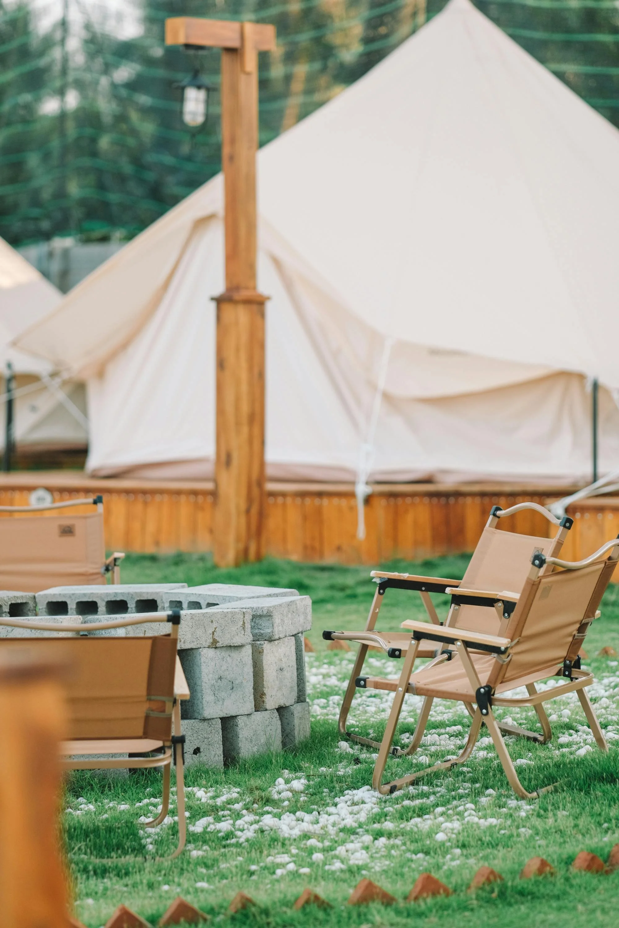 Outdoor camping scene with beige camping chairs, a fire pit made of concrete blocks, a wooden post, and a large beige canvas tent.