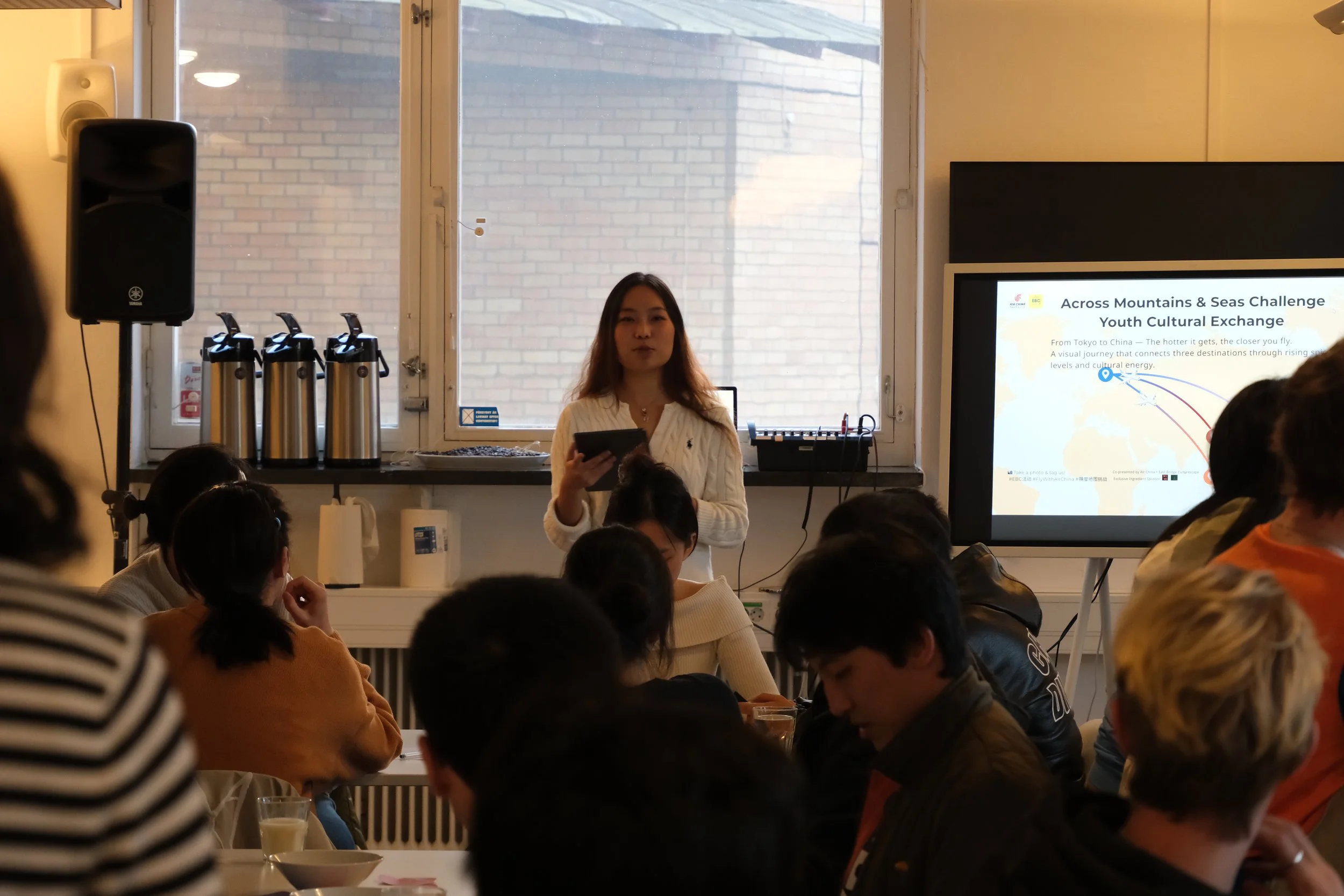 A woman giving a presentation to a group of people in a room with a large window and a screen displaying a slide about cross-cultural exchange.