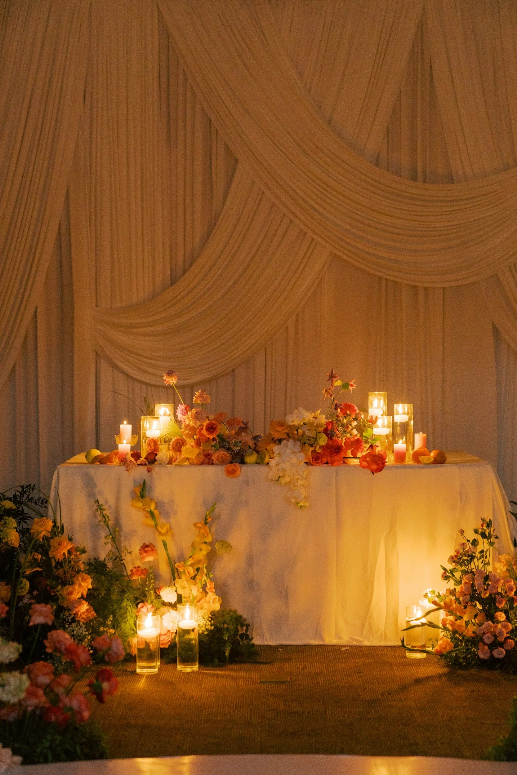 A table decorated with flowers and lit candles, set against a backdrop of white draped fabric.