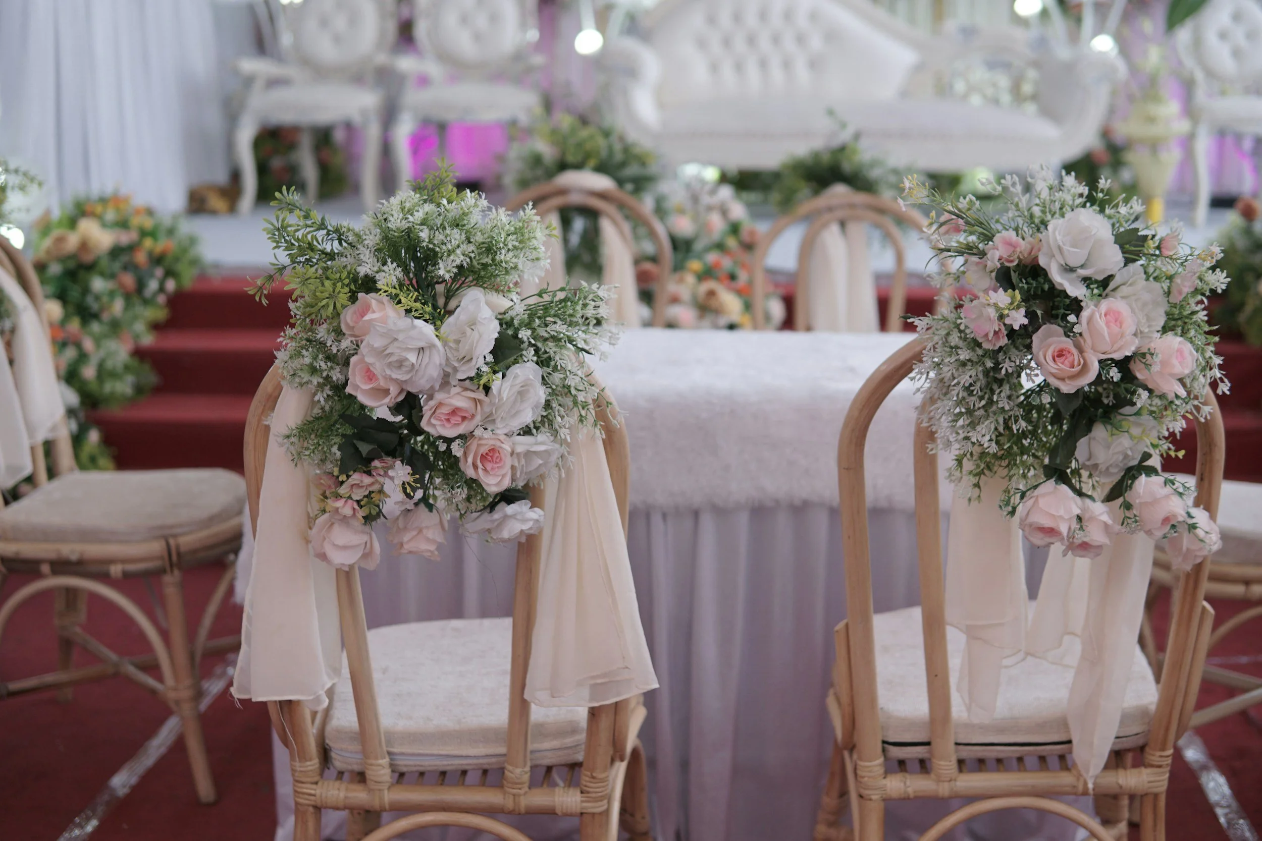 Decorated wedding chairs with white drapes and pink and white floral arrangements at a wedding ceremony with a decorated stage in the background.