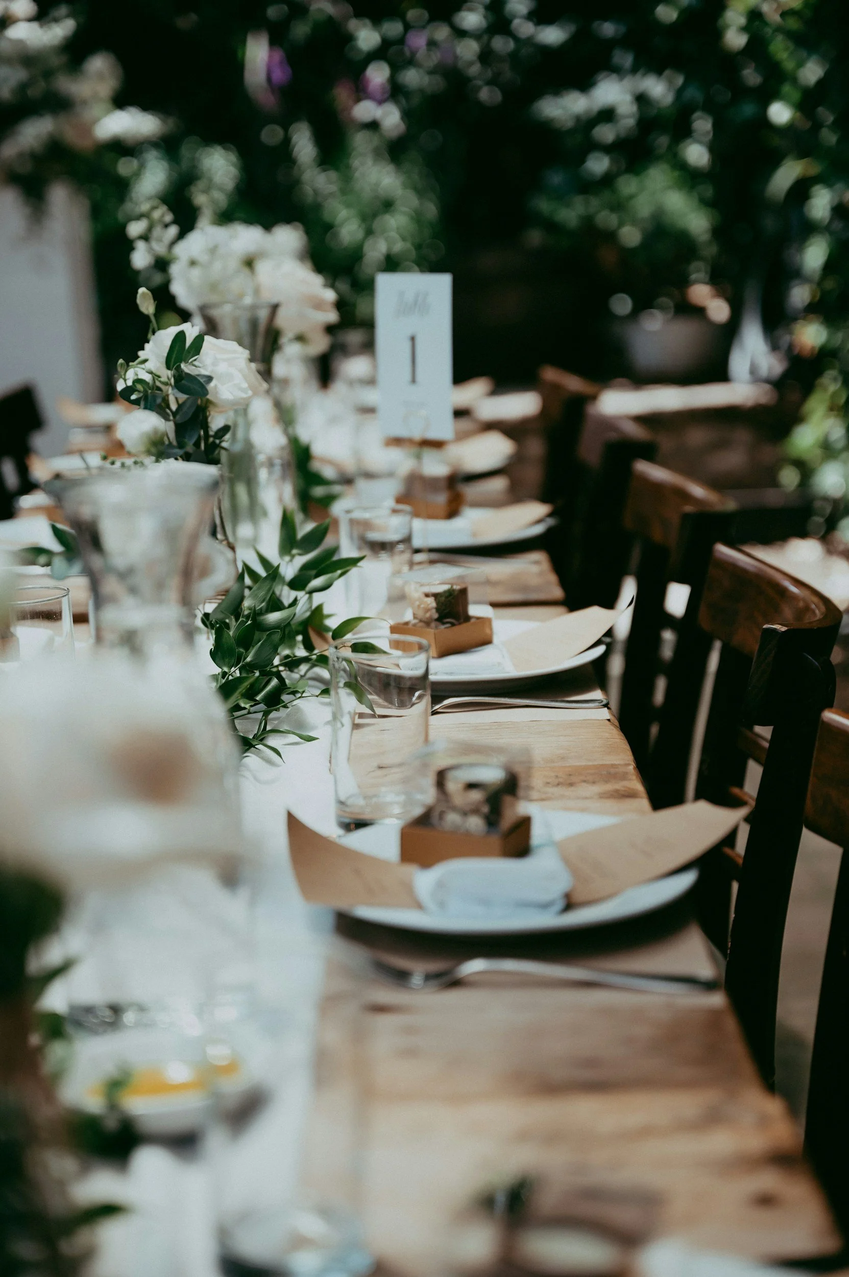 A wedding reception table decorated with white flowers, greenery, candles, and tableware, set outdoors during daytime.