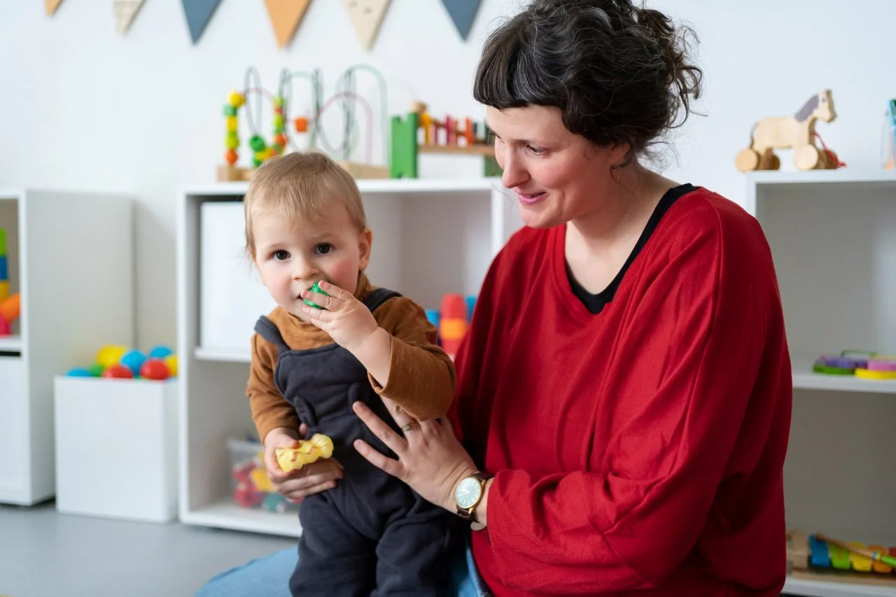 A woman holding a young child in a playroom with colorful toys and decorations, the child eating a cookie.