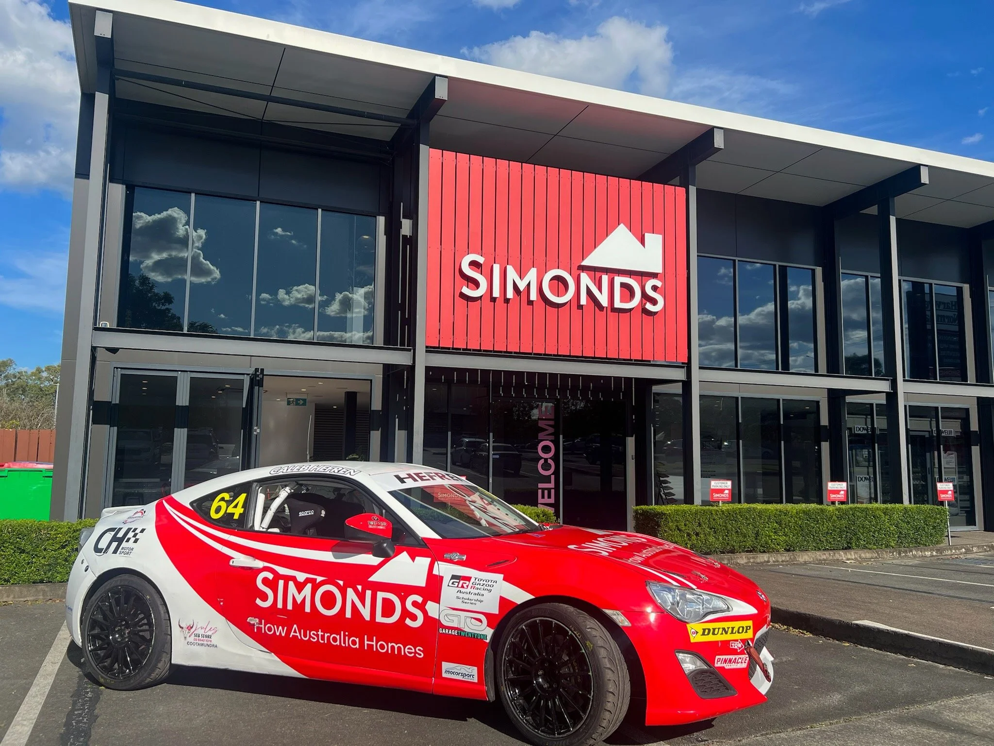 A red and white racing car parked in front of a modern store with a large red sign that reads 'SIMONDS' and a mountain logo, reflecting the sky in its windows.