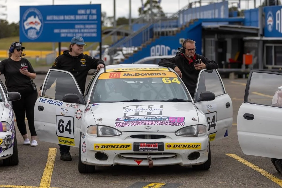 Caleb Hefren's car with number 64 and McDonald's branding parked in a pit lane at a racing event with three team members around it, one looking at a phone, one holding the car door, and another looking at a device.