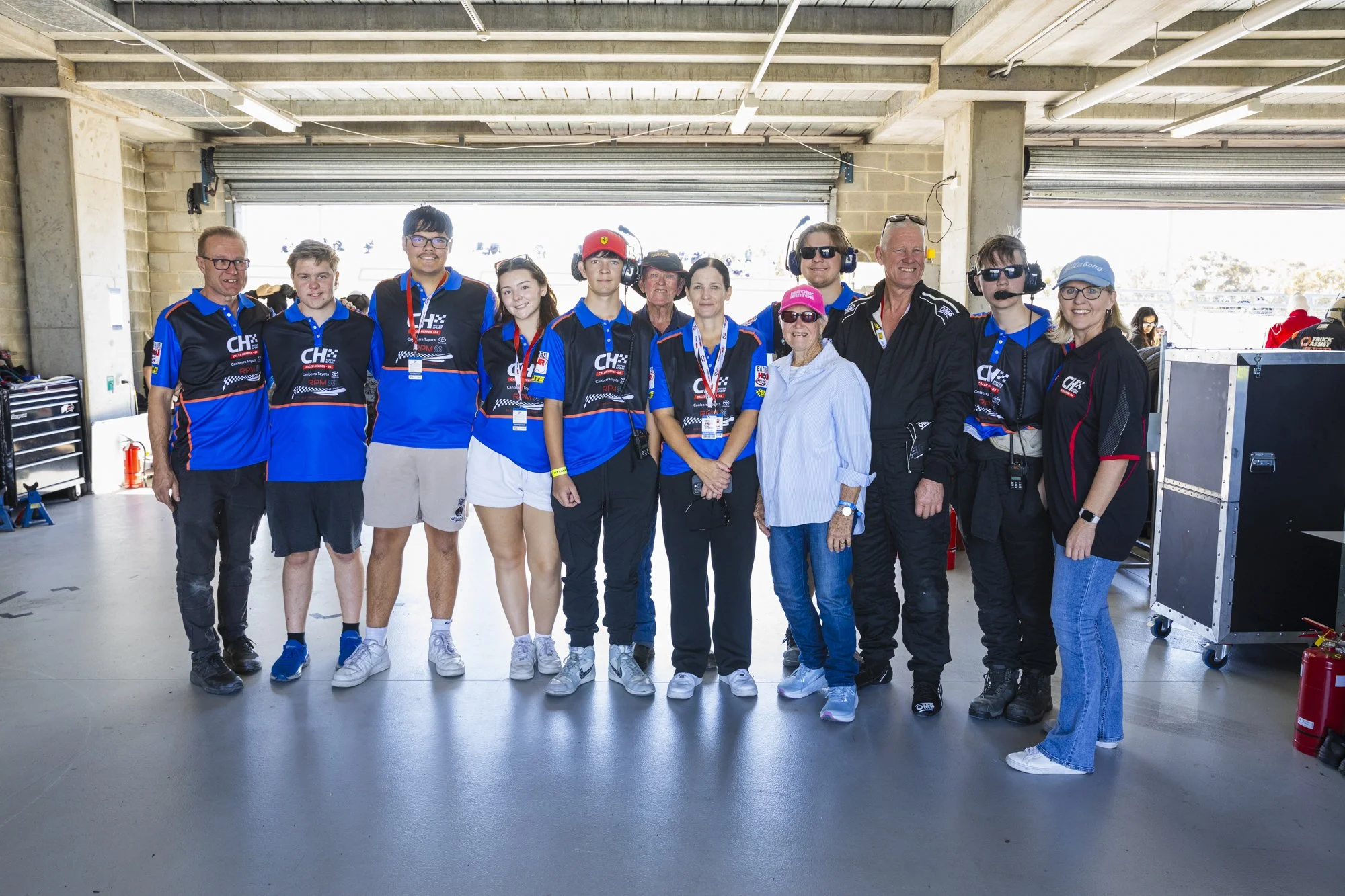 Group of racing team members and officials standing together in garage area at a racetrack, wearing team uniforms and headsets.