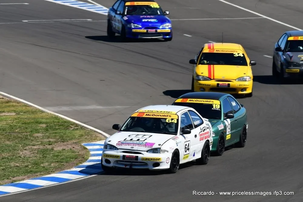 Series of race cars on a track, including a white car with a large hood decal leading, followed by a green and black car, a yellow car, and a blue car behind, all racing on a curved section of the track.
