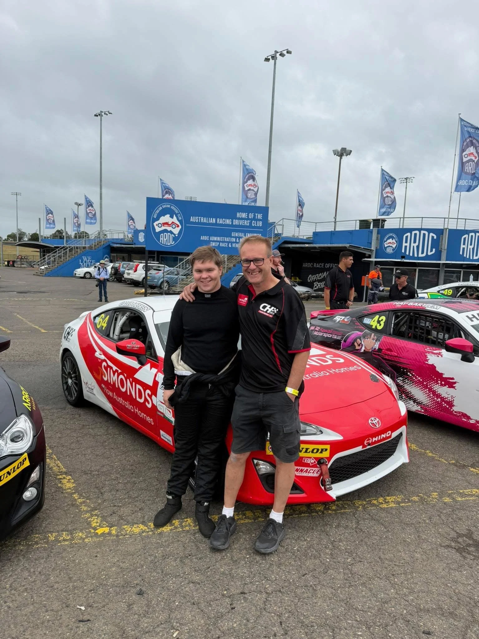 Two men - Caleb and Dean Hefren - standing in front of a race car at an Australian racing event, smiling and posing for a photo.