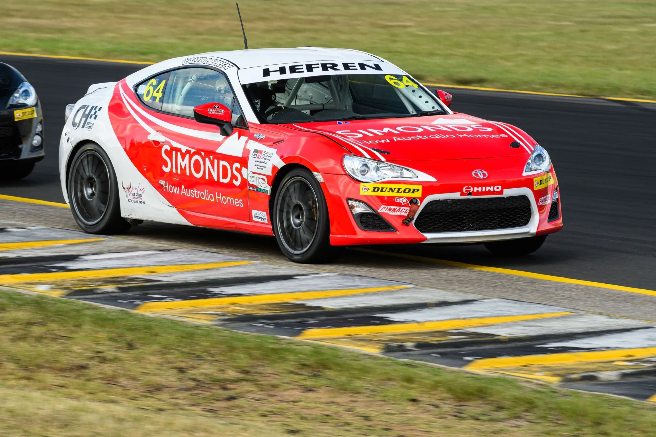 A red and white Toyota sports car with racing sponsors, number 64, on a race track during a race.