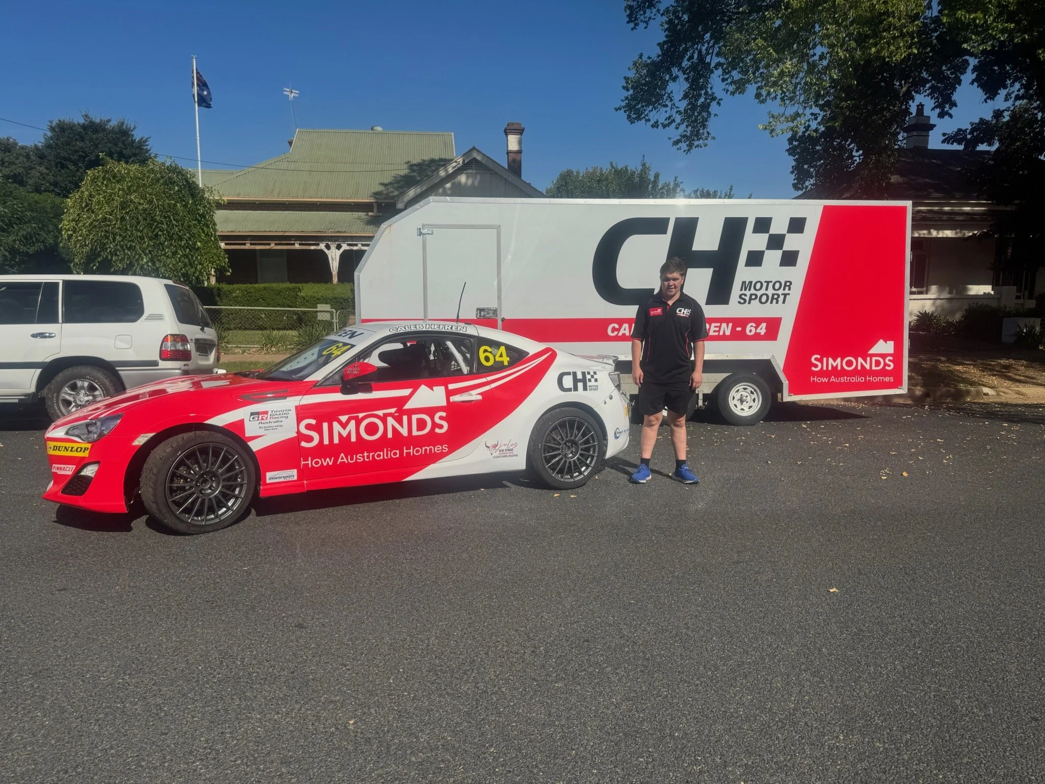 A race car with red and white livery and the number 64 on its windshield, displaying suit sponsor 'Simonds How Australia Homes', is parked next to a large white trailer with matching branding. A man in a black racing team shirt and shorts is standing