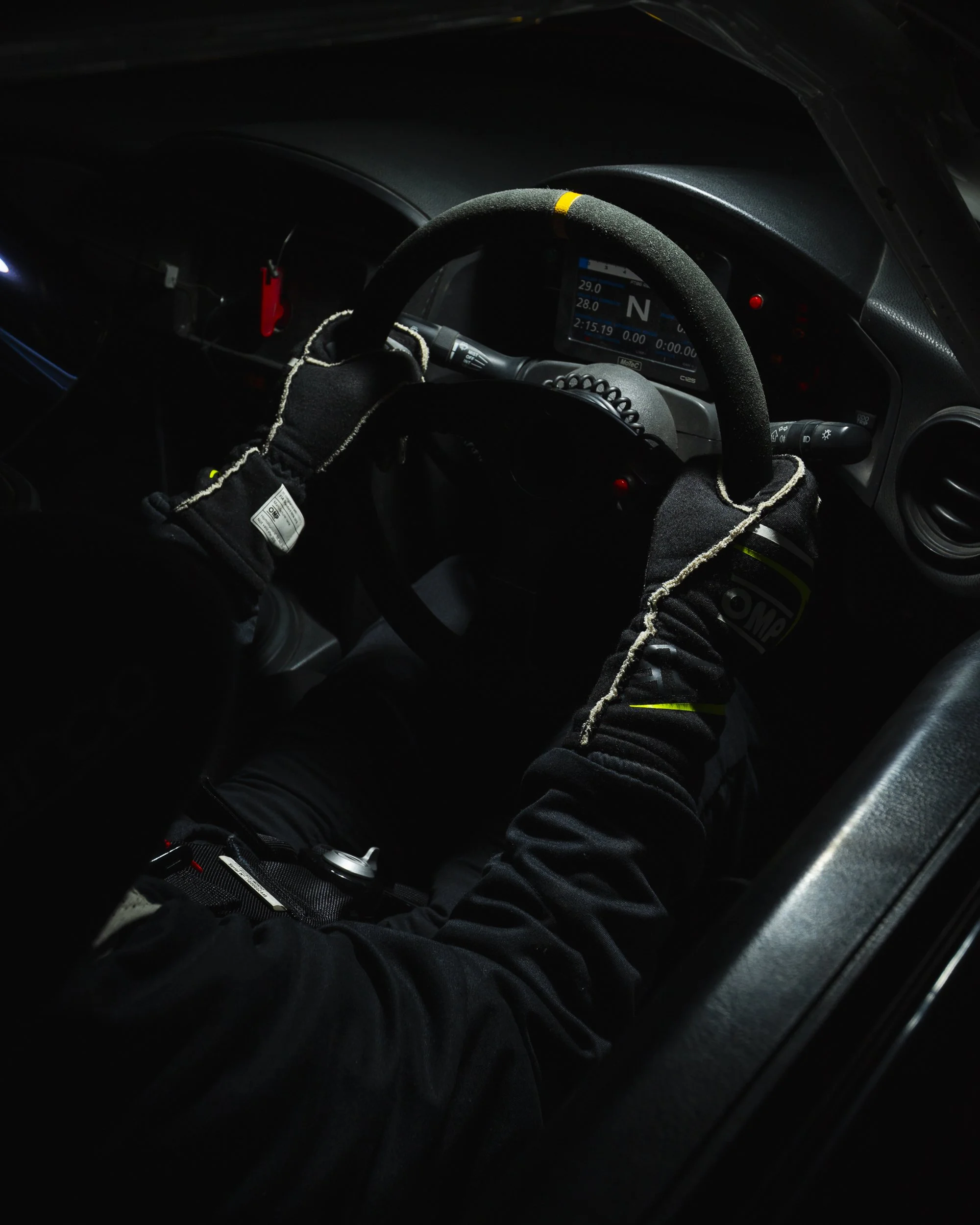 Close-up of a race car driver in a helmet and racing suit gripping a steering wheel inside a race car cockpit.
