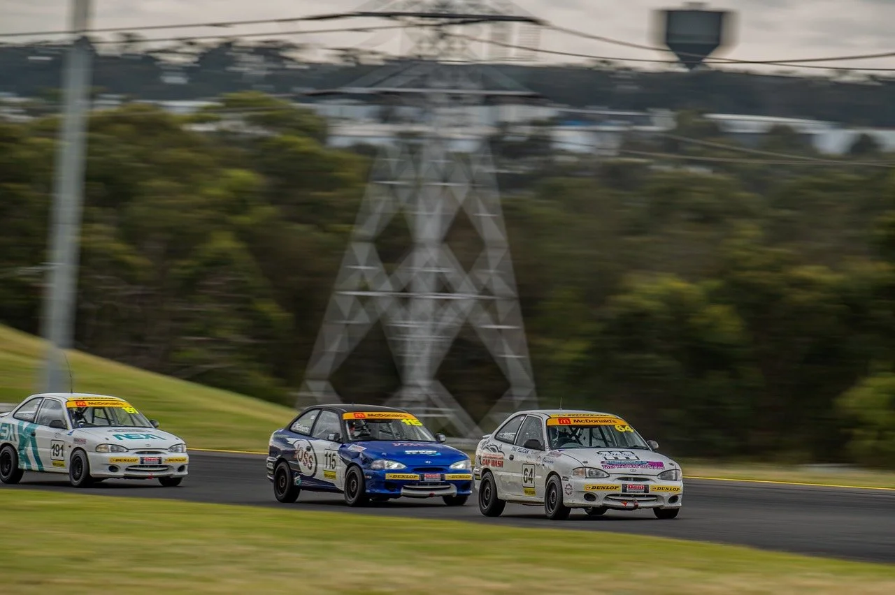 Three race cars on a track, racing side by side in a motorsport event, with a blurred background of trees and electrical transmission towers.