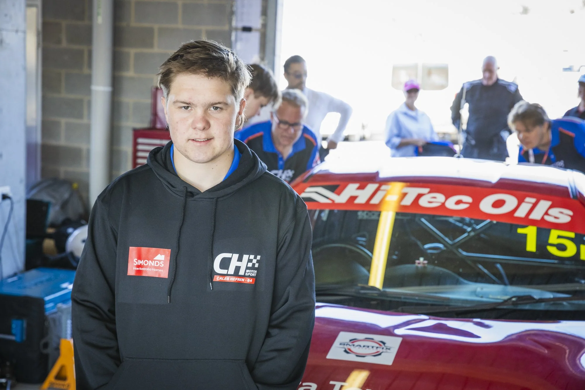 Young male race car driver standing in garage with race car and team members in background