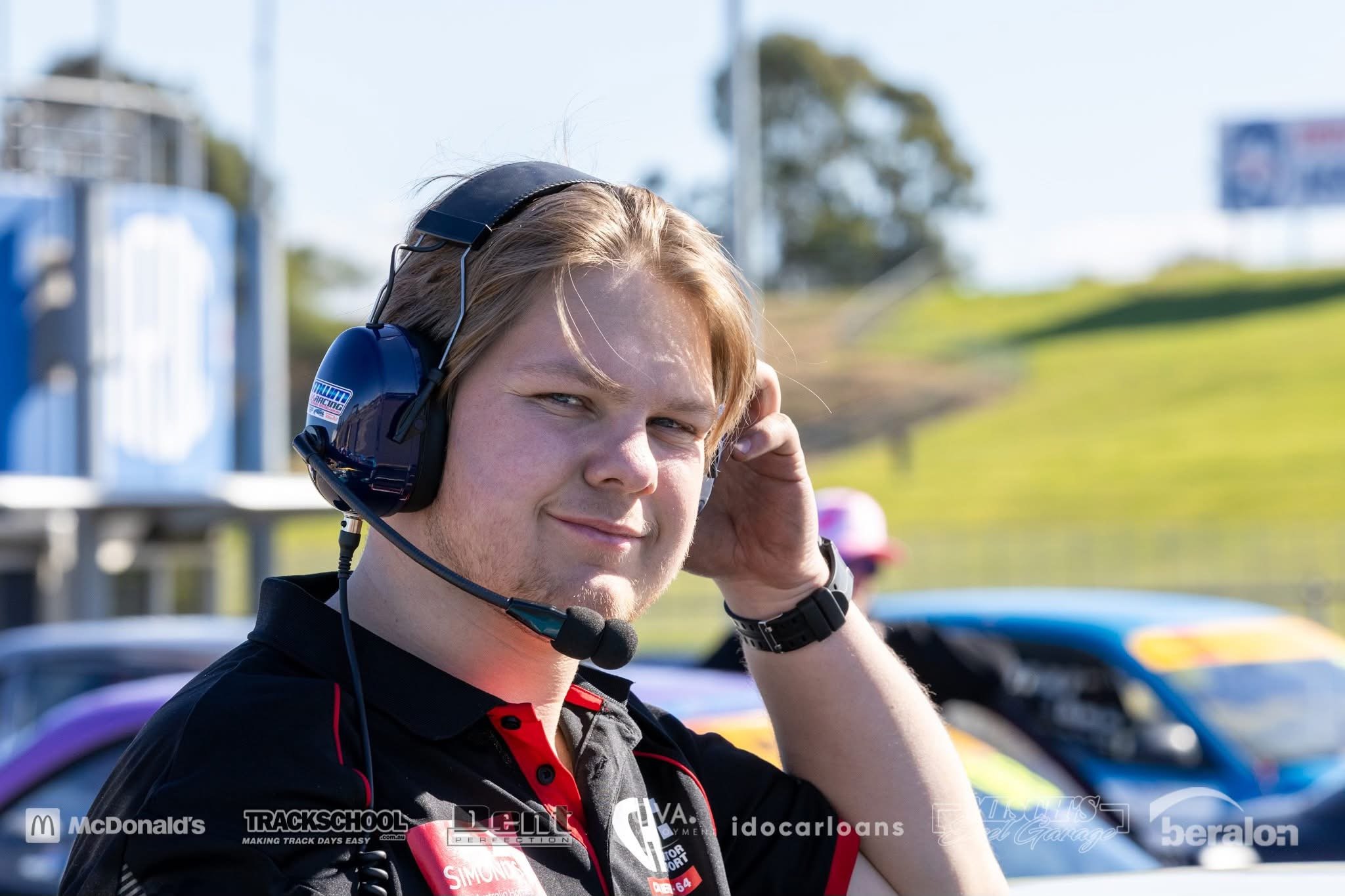 Young man wearing blue headphones with microphone, blue and black sports jacket, standing under bridge or overpass, smiling.