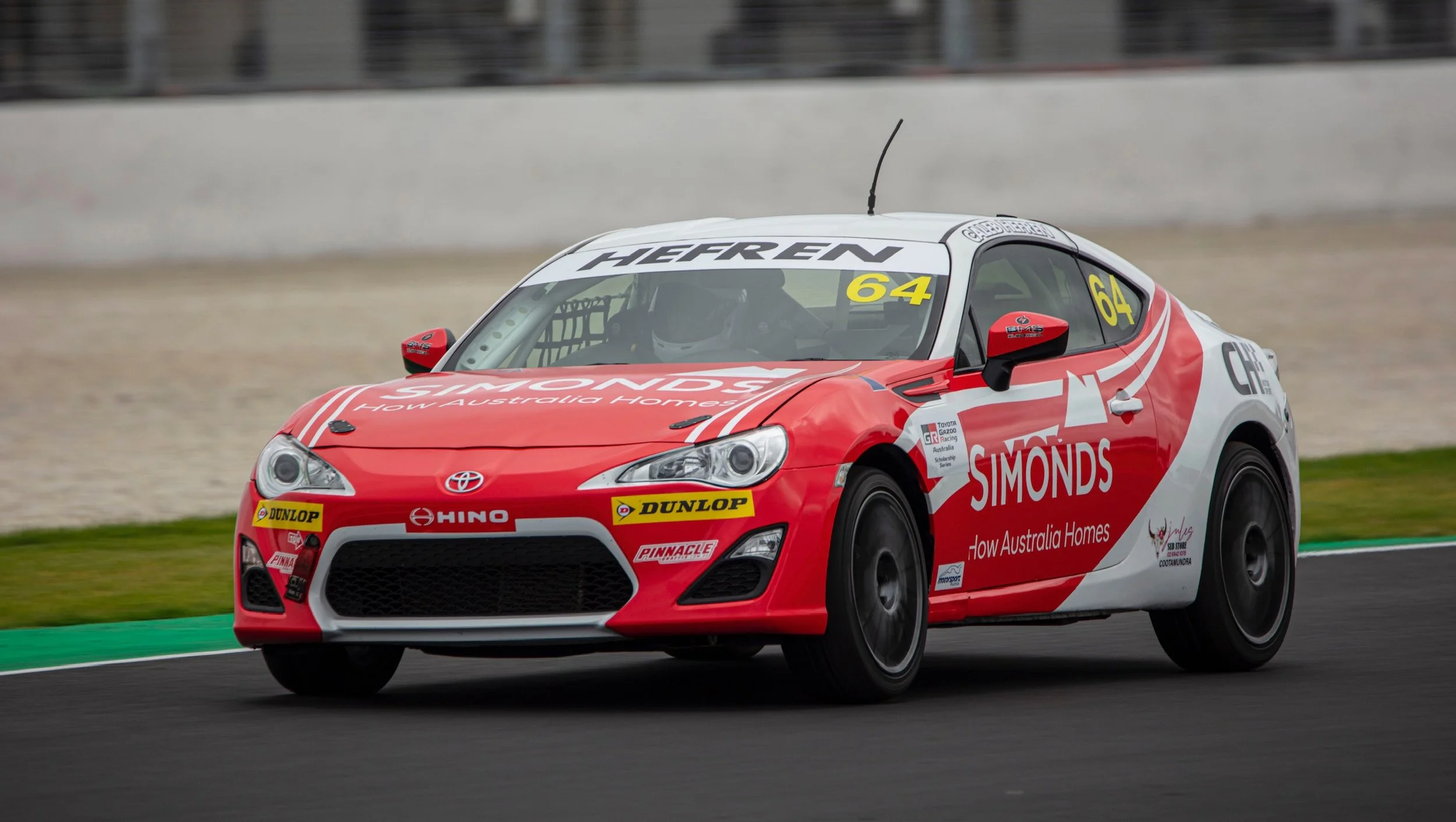 A race car driving on a track, white with red accents, displaying sponsor logos including Simonds and Dunlop.
